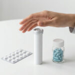 Une main au-dessus d'un tube de médicaments à côté d'une plaquette de comprimés et d'un flacon de gélules bleues.