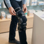 Une personne ajuste une genouillère articulée orthopédique dans une pharmacie avec des étagères de boîtes médicales.