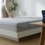 Une personne touche un matelas médical moderne dans une chambre lumineuse avec une tablette affichant des données financières.