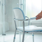 Main posée sur un siège de douche ergonomique avec structure métallique dans une salle de bain moderne.