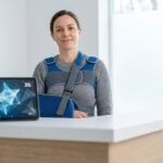 Femme portant un gilet d'immobilisation d'épaule dans un cabinet médical avec une tablette affichant des données.
