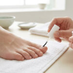 Mains appliquant un vernis de traitement sur l'ongle d'un pied posé sur une serviette blanche.