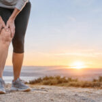 Une personne debout sur un sentier tient son genou douloureux au coucher du soleil, symbolisant l'arthrose.