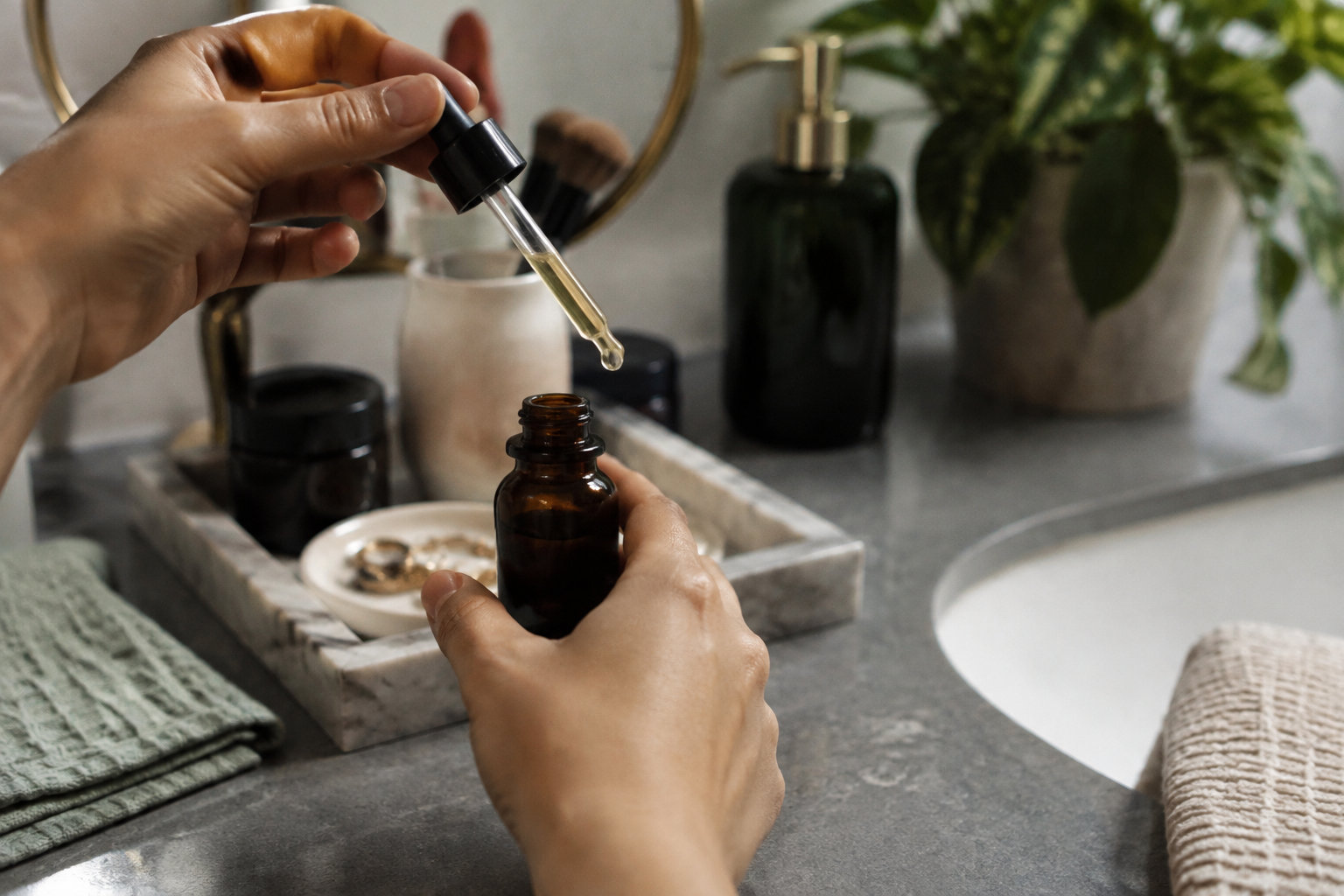 Mains tenant une pipette de sérum au-dessus d'un flacon en verre ambré dans une salle de bain lumineuse.