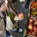 Une personne choisit une pomme fraîche sur un étal de marché avec un sac en filet rempli de légumes.