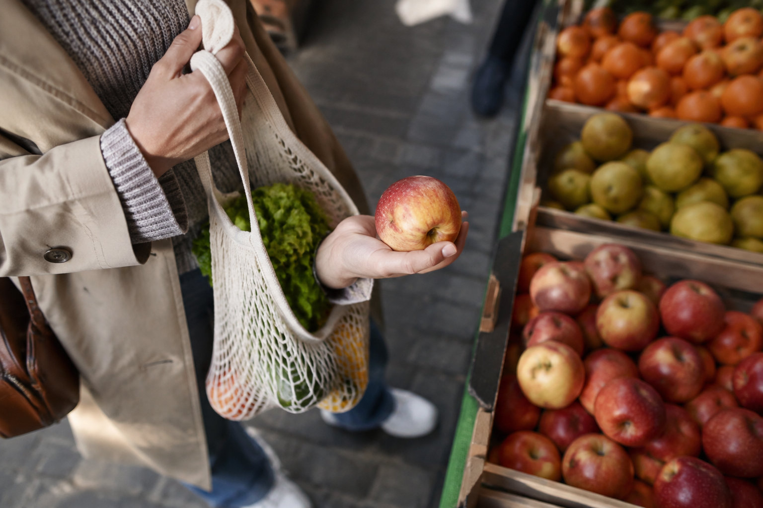 Une personne choisit une pomme fraîche sur un étal de marché avec un sac en filet rempli de légumes.