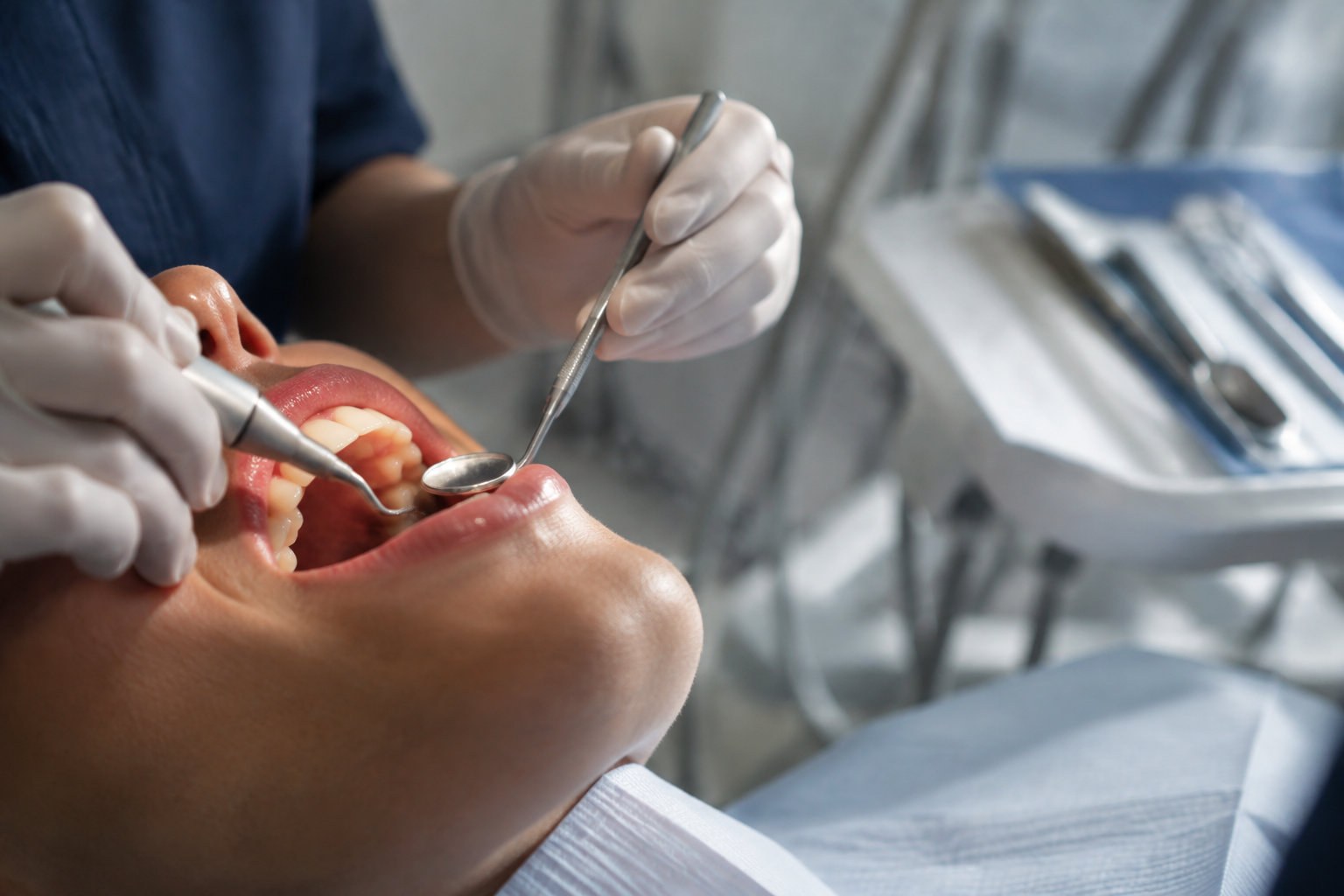 Un dentiste en gants procède à un détartrage avec un miroir et un instrument ultrasonique sur un patient allongé.