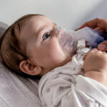 Un bébé allongé sur le dos recevant un traitement par aérosol via un masque respiratoire tenu par une main d'adulte.
