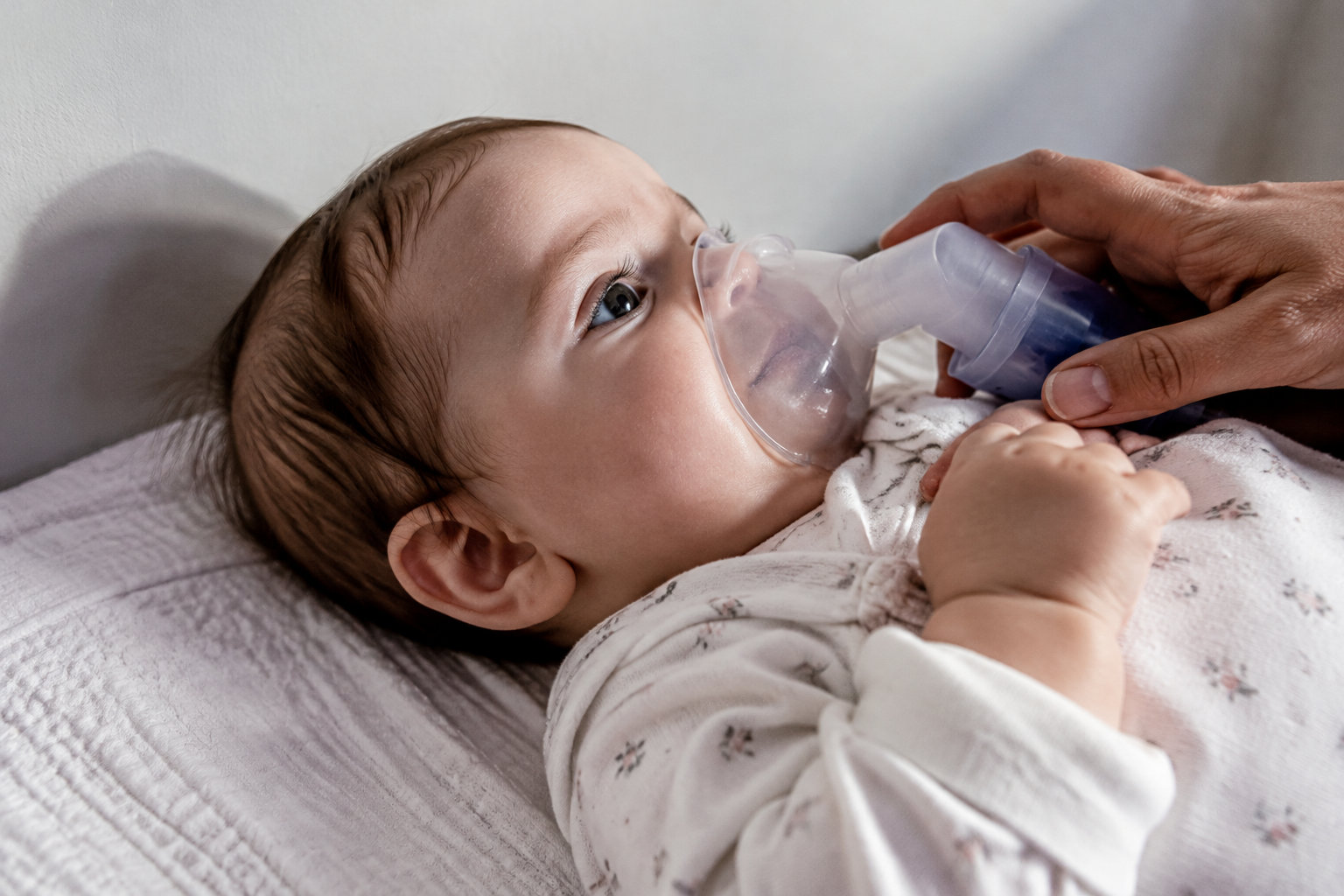 Un bébé allongé sur le dos recevant un traitement par aérosol via un masque respiratoire tenu par une main d'adulte.
