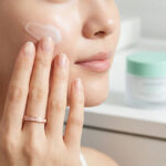 Une femme s'applique une crème hydratante blanche sur la joue, avec un pot de soin flouté en arrière-plan.